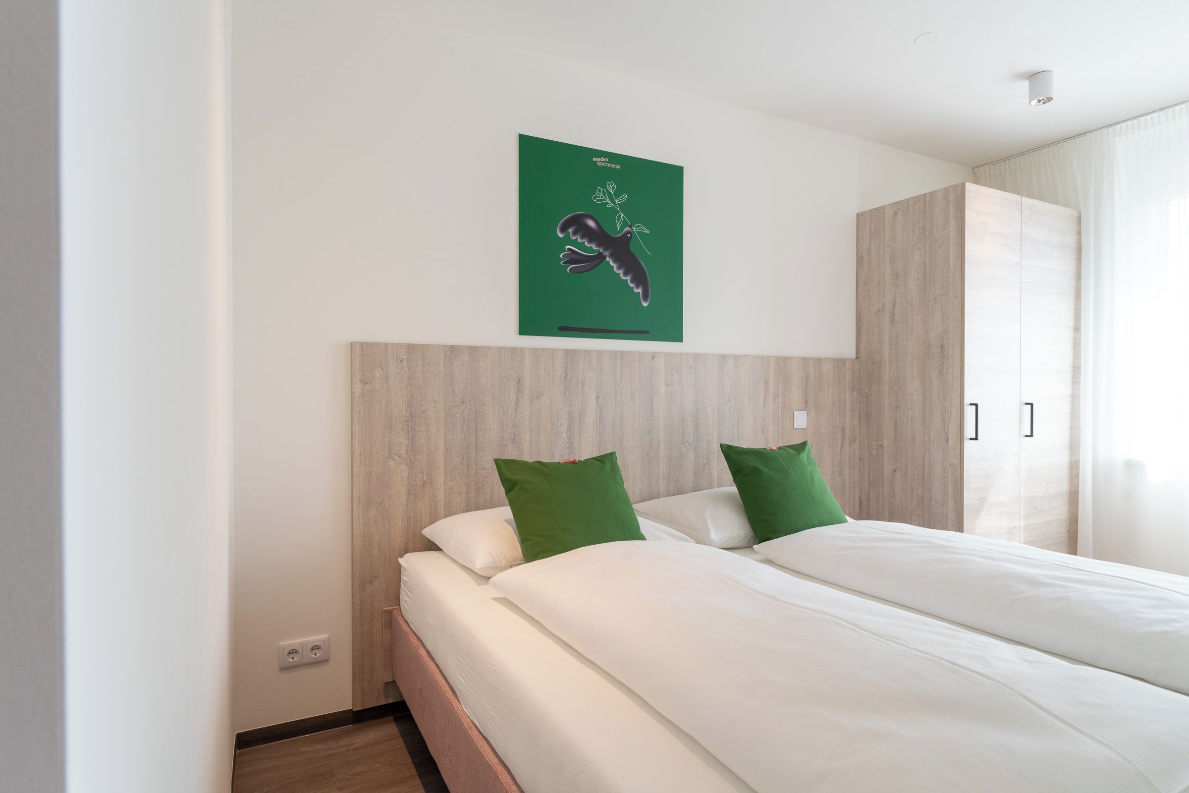 Una habitación de hotel con cama, almohadas verdes, cabecera de madera, cuadro con motivos verdes y una ventana con cortinas.