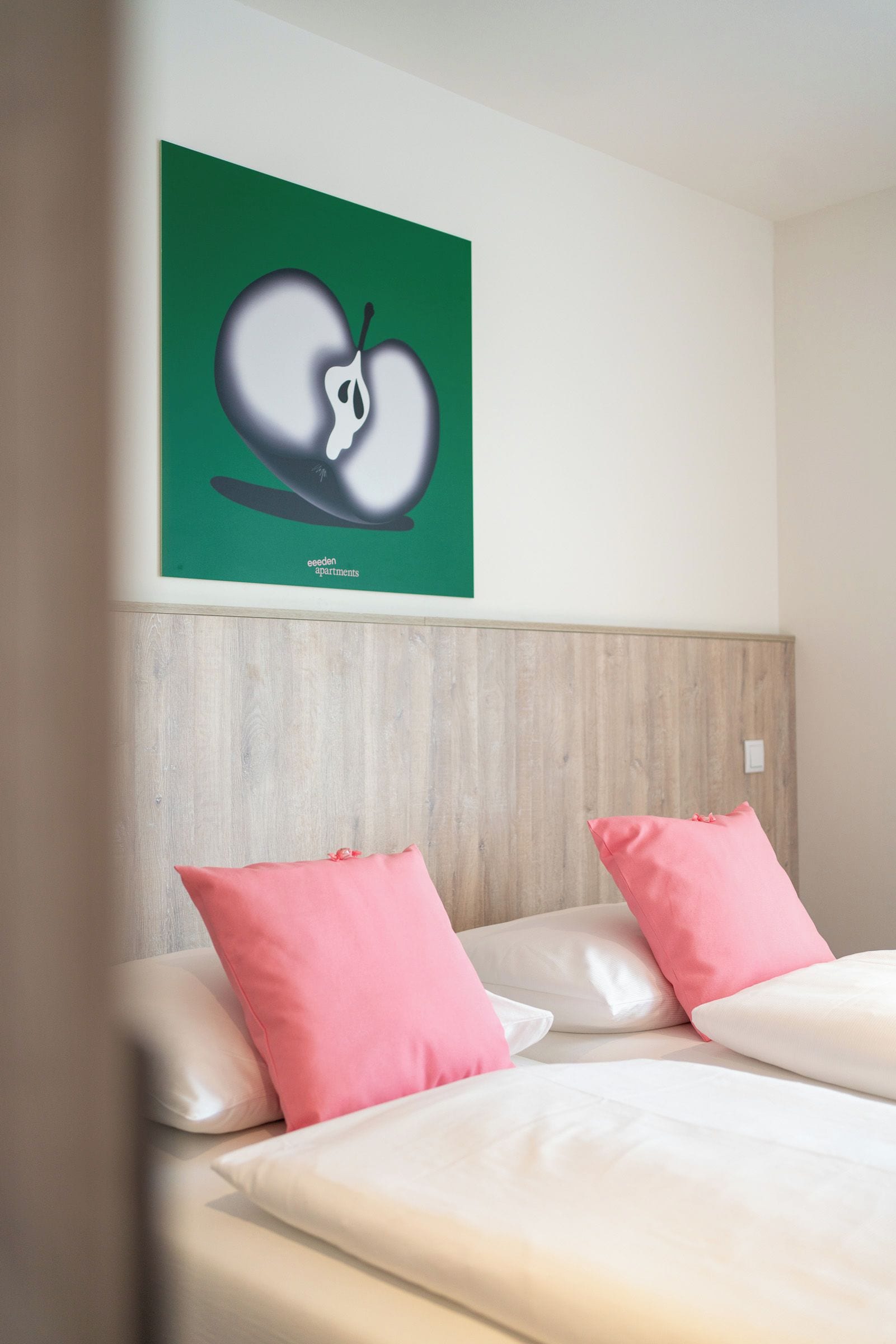 Une chambre d’hôtel avec un lit, deux oreillers roses, un mur vert orné d’une illustration de fruits et une tête de lit en bois.