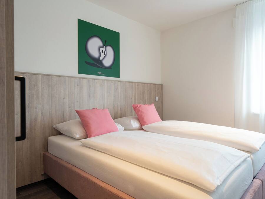 Cama doble con dos almohadas, cabecero de madera y pared verde con una obra de arte en forma de corazón.