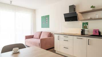 A modern kitchenette with a coffee machine, a small shelf with glassware, and a dining area with a table and a pink sofa.