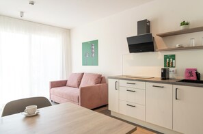 Una kitchenette moderna con cafetera, un estante pequeño con cristalería y un comedor con mesa y sofá rosa.