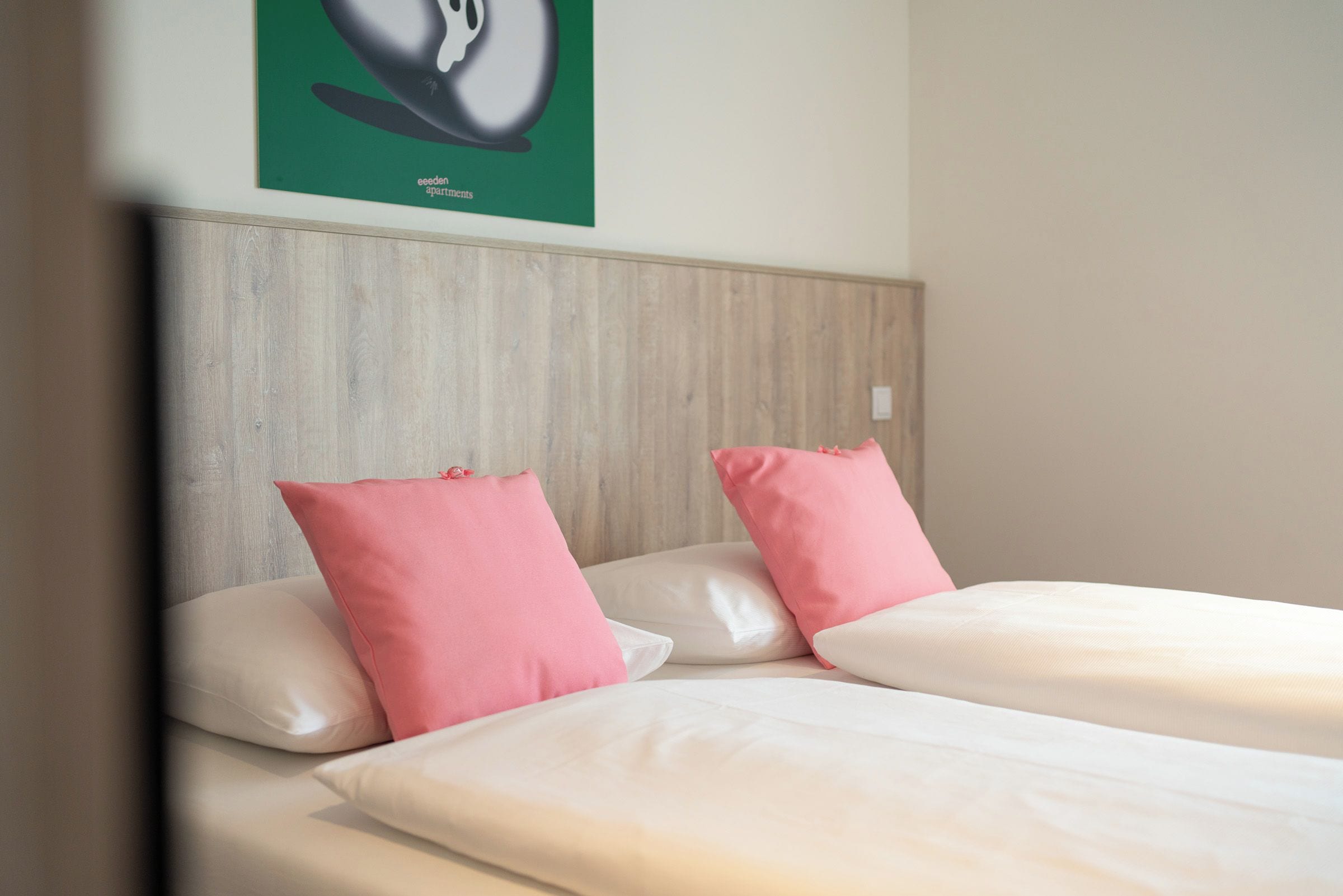 Quarto de hotel com cabeceira de madeira, duas camas com almofadas brancas e cor-de-rosa, e um quadro verde na parede.