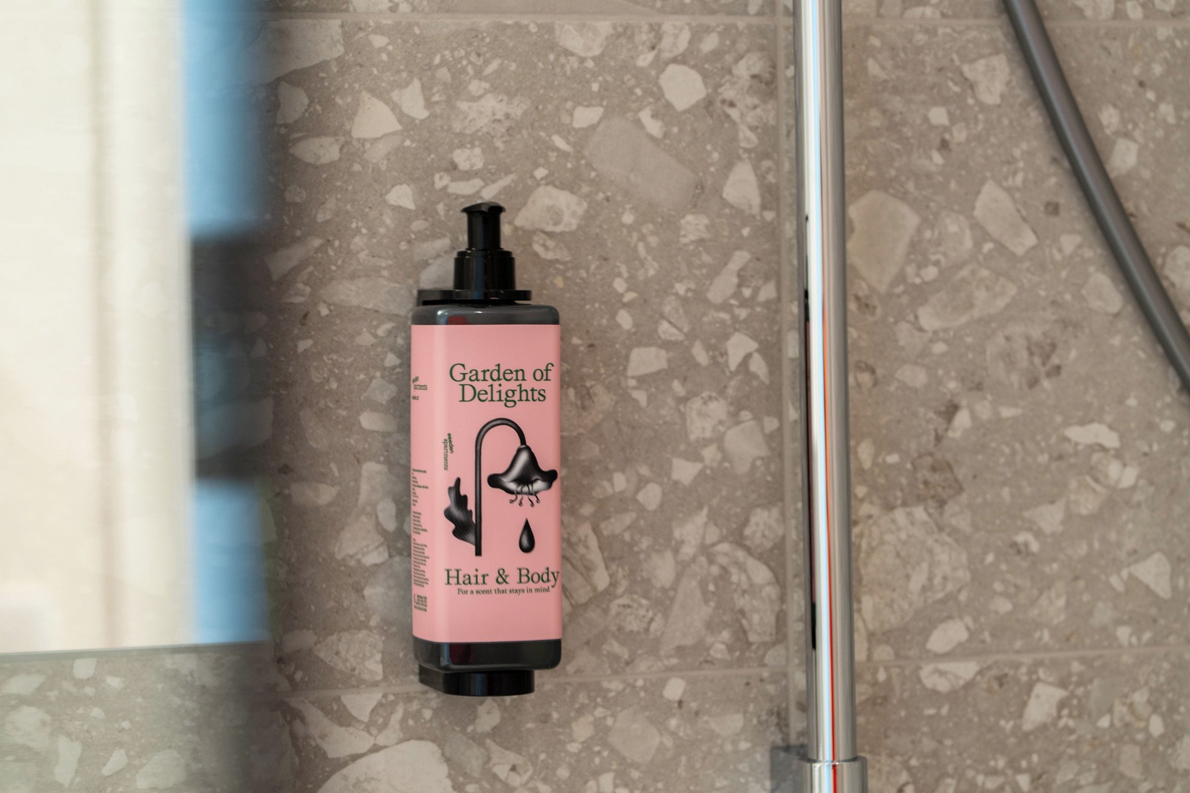 A bathroom with a pink bottle labeled 'Garden of Delights Hair & Body' on a speckled tile floor.
