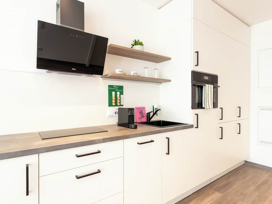 A modern kitchen with white cabinetry, a black range hood, a built-in oven, and a countertop with a kettle and a coffee maker.