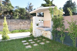 A stone barbecue area with a metal grill, surrounded by a stone wall and a grassy lawn with stepping stones. - Mon Emvasia Margarites Villa B (Rethymno)