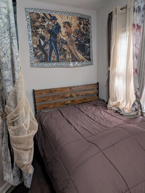 A bedroom with a bed, a wooden headboard, a wall painting, and curtains. - 146-5       Herbie Hancock, Pianist Bask!                       1Bedroom Studio. (New London)