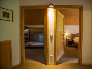 Ein Zimmer mit einer Holztür, die zu einem Schlafbereich mit Etagenbetten führt. Ein kleiner Nachttisch mit einer Lampe und einem gerahmten Bild sind vorhanden.