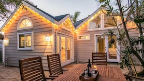 En terrasse med træmøbler og et spisebord, omgivet af et hus med udendørs belysning.