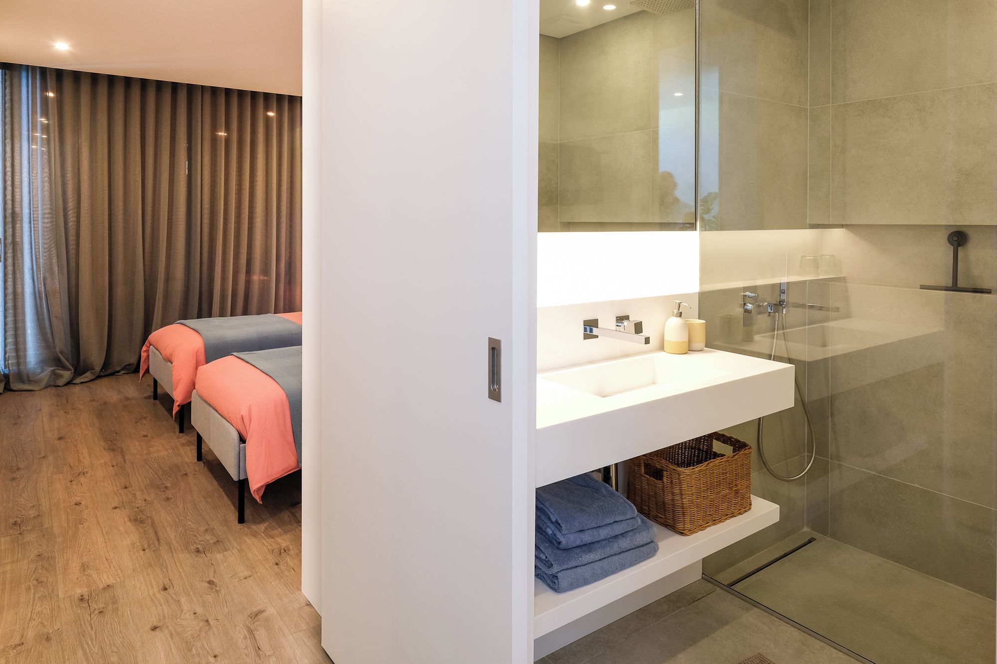 A modern bathroom with a glass-enclosed shower, a white vanity with a mirror, and a bed with orange and blue bedding in the adjacent room.