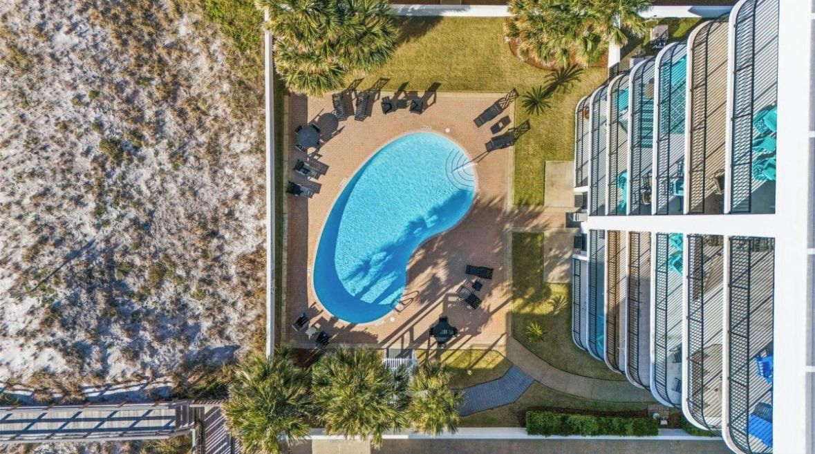 Vue aérienne d’un hôtel doté d’une piscine entourée de chaises longues et de parasols, situé à côté d’un immeuble avec des balcons.