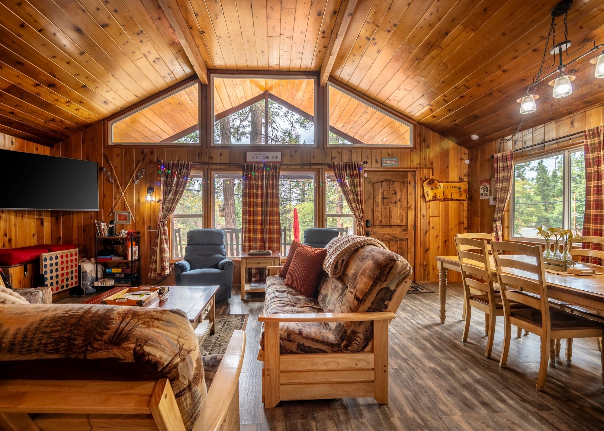 A cozy cabin interior with a wooden ceiling, a large window, a dining table, and a sofa.