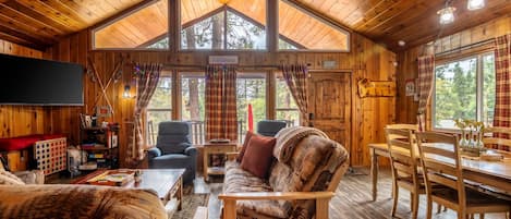 A cozy cabin interior with a wooden ceiling, a large window, a dining table, and a sofa.