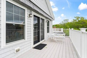 Eine weiße Holzdielen-Terrasse mit weißer Geländer, einer schwarzen Tür und einem Fenster mit Rollläden.