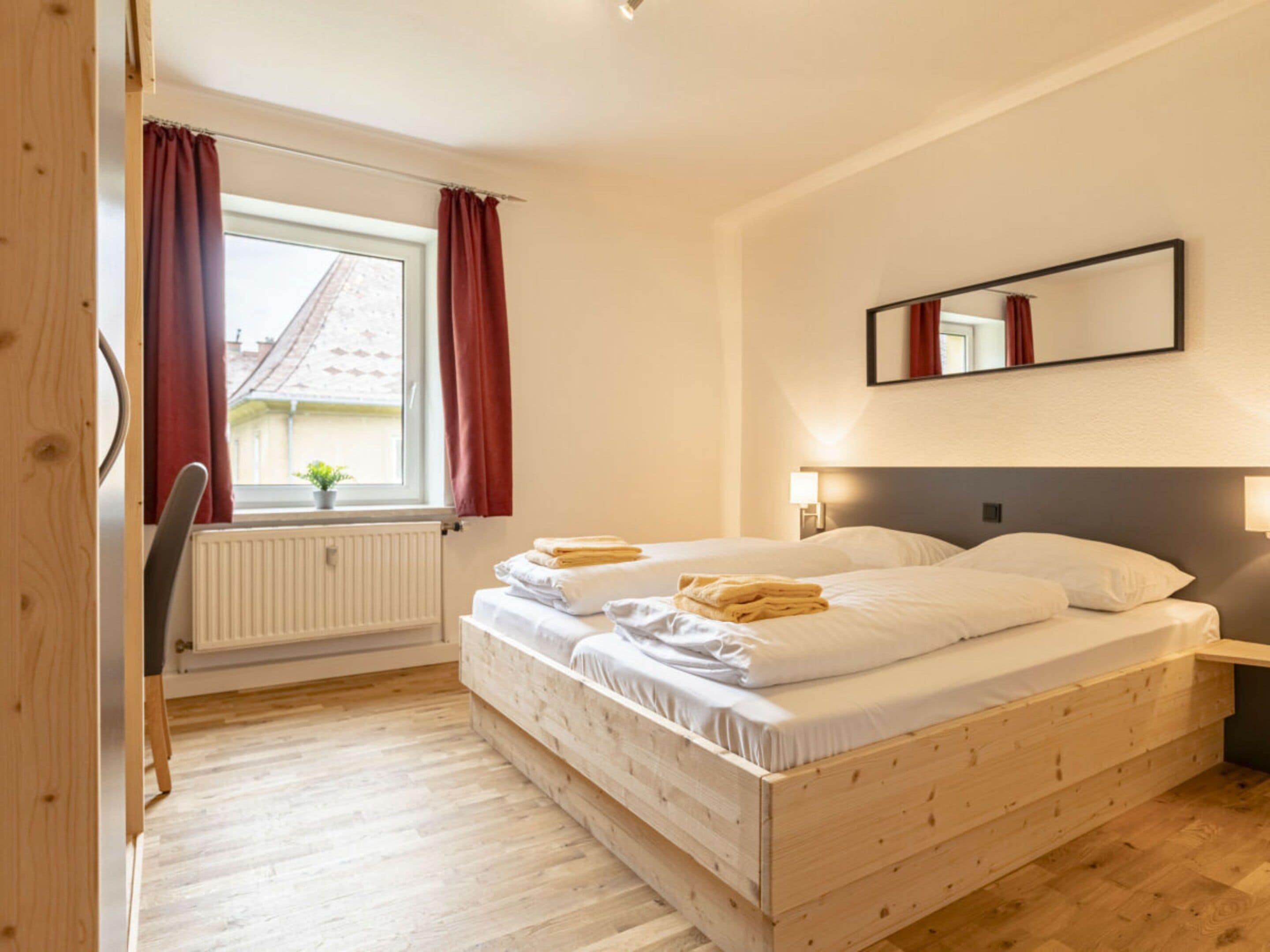 A double room with a wooden bed, a window with curtains, a radiator, and a small plant on the windowsill.