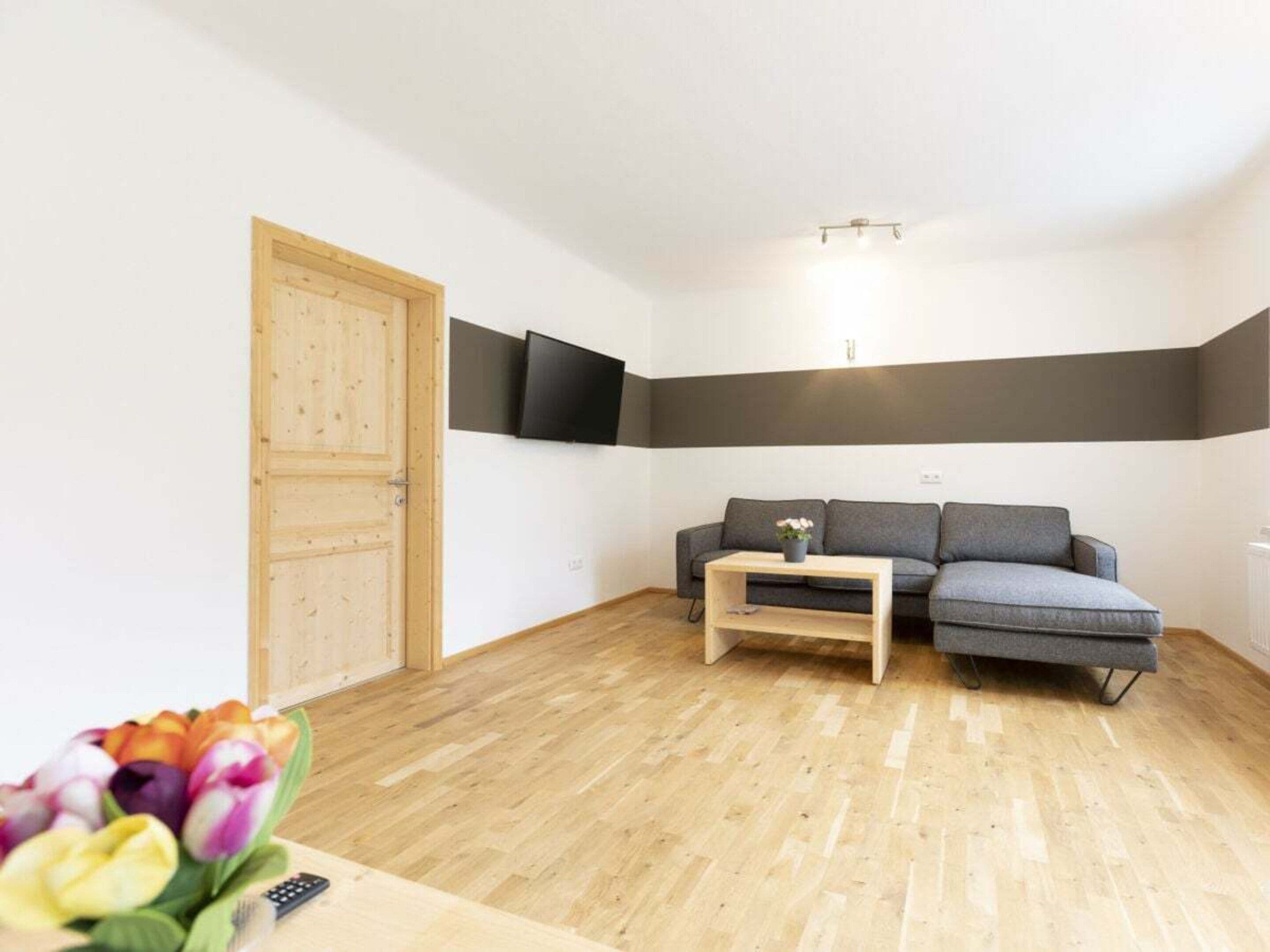 A room with a wooden door, a grey sofa, a small wooden table, and a flat-screen TV mounted on the wall.