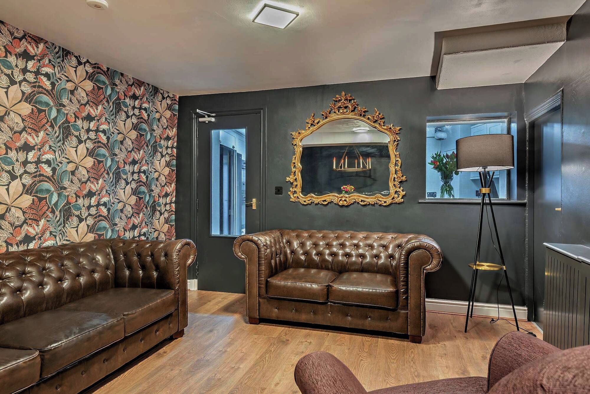 Une pièce avec un mur de couleur sombre, un papier peint à motifs floraux, un grand miroir, un canapé en cuir marron et un lampadaire.