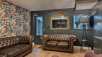 Une pièce avec un mur de couleur sombre, un papier peint à motifs floraux, un grand miroir, un canapé en cuir marron et un lampadaire.