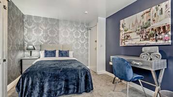 A bedroom with a bed, a desk, a chair, a painting, and a patterned wallpaper.
