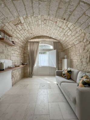 A room with a stone ceiling, a sofa with patterned cushions, a wooden shelf with kitchenware, and a window with curtains. - Vrbo Property (Locorotondo)