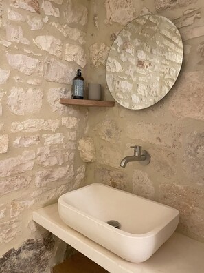 A bathroom with a stone wall, a round mirror, a wooden shelf with toiletries, and a white sink. - Vrbo Property (Locorotondo)