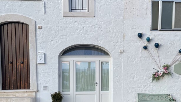 Um edifício branco com porta marrom, uma janela com grade de metal e um pequeno vaso com planta.