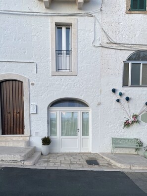 A white building with a brown door, a window with a metal grille, and a small potted plant. - Vrbo Property (Locorotondo)