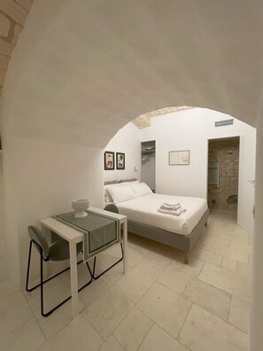 Ein Schlafzimmer mit einem Bett, einem kleinen Tisch und einem Stuhl. Ein Durchgang führt in einen anderen Raum.