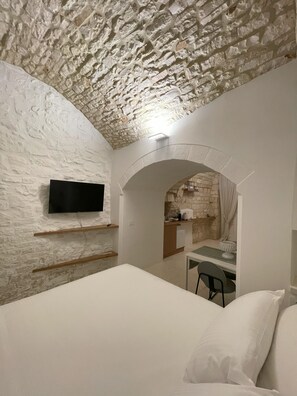 A bedroom with a stone wall, a flat-screen TV mounted on the wall, a bed with white bedding, and a small table with a chair. - Vrbo Property (Locorotondo)