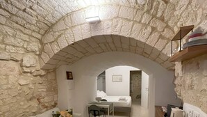 Um quarto aconchegante com parede de pedra, um sofá branco com almofadas estampadas, uma pequena mesa de jantar e um espaço com cozinha compacta.