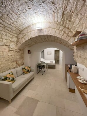 A cozy room with a stone wall, a white sofa with patterned cushions, a small dining table, and a kitchenette area. - Vrbo Property (Locorotondo)