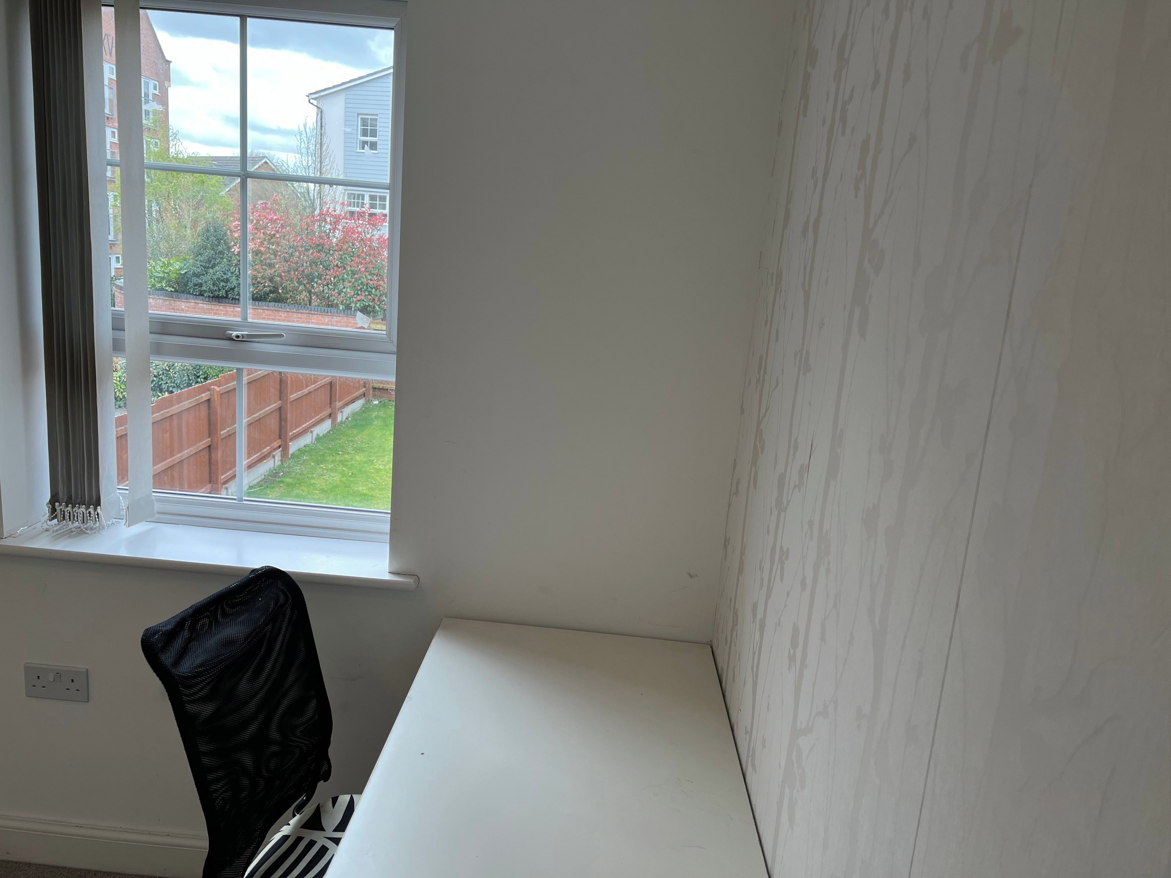 Une pièce avec un bureau blanc, une fenêtre donnant sur un jardin et un bâtiment, et un radiateur fixé au mur.