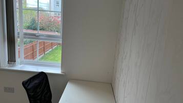 A room with a white desk, a window with a view of a garden and a building, and a radiator on the wall.