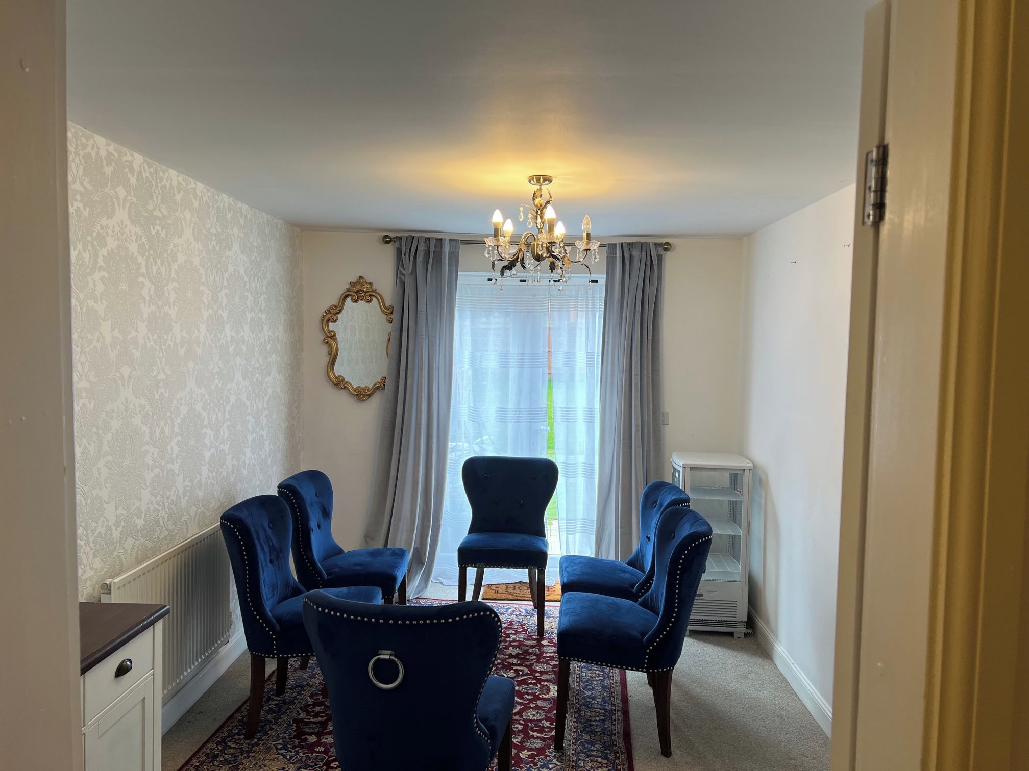 A room with a chandelier, a large mirror, two blue chairs, a radiator, and a window with curtains.
