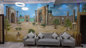 A room with a mural of a historic building, a grey sofa set, and a chandelier. - Hotel Dilkash (Samarkand)