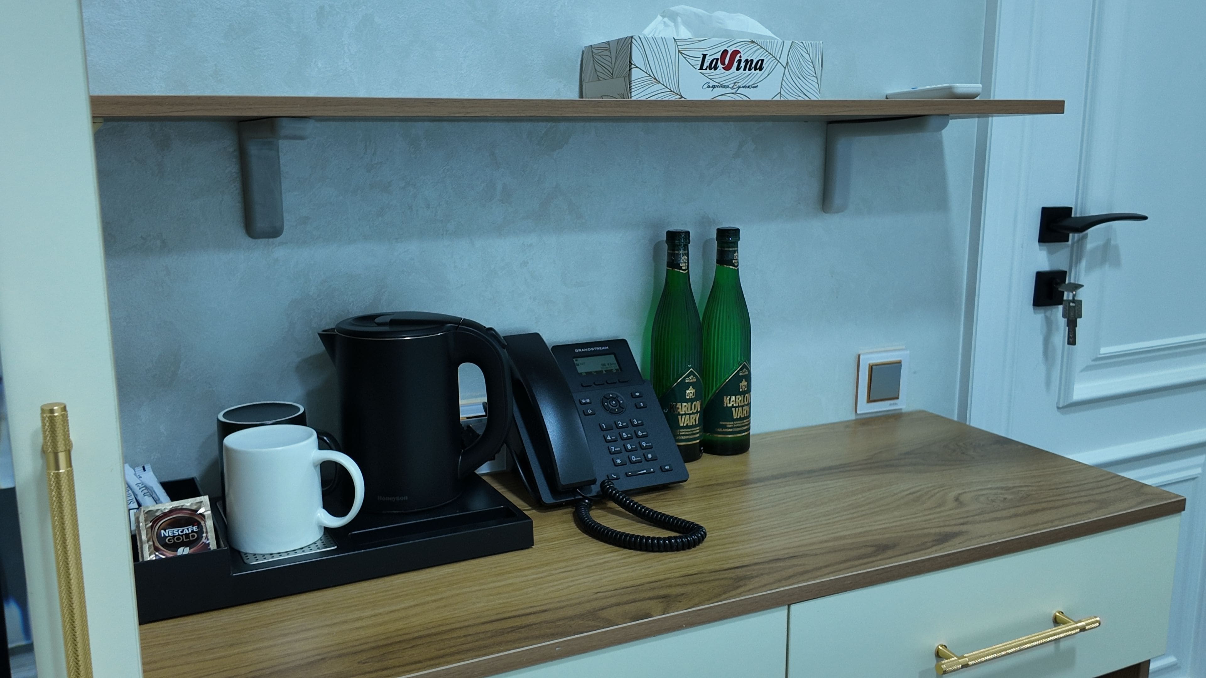 Habitación de hotel con cafetera, teléfono, taza de café y dos botellas de agua sobre una encimera de madera.