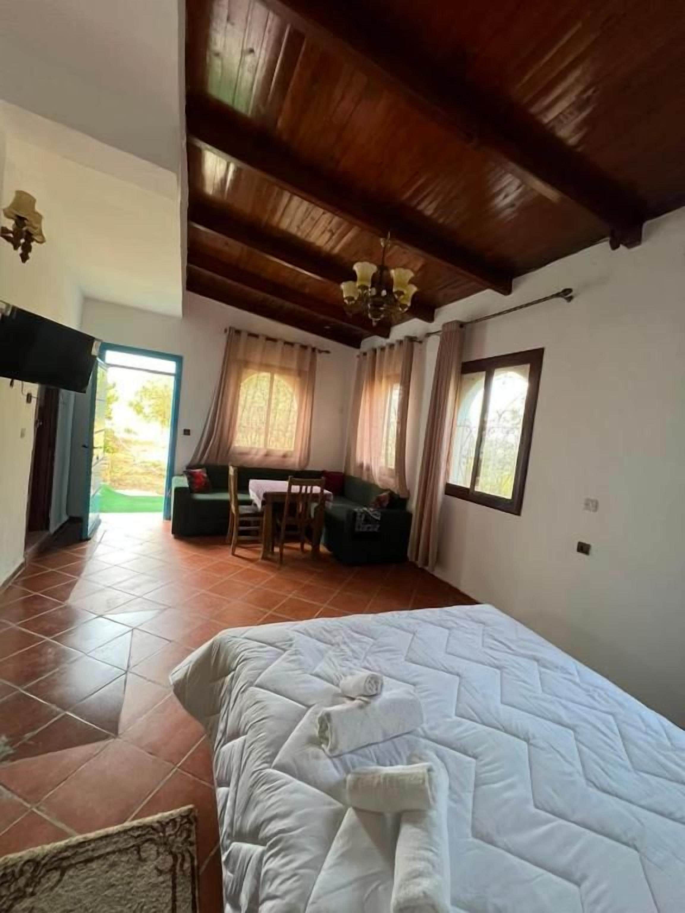 A bedroom with a large bed, wooden ceiling beams, a desk with a chair, and a window.