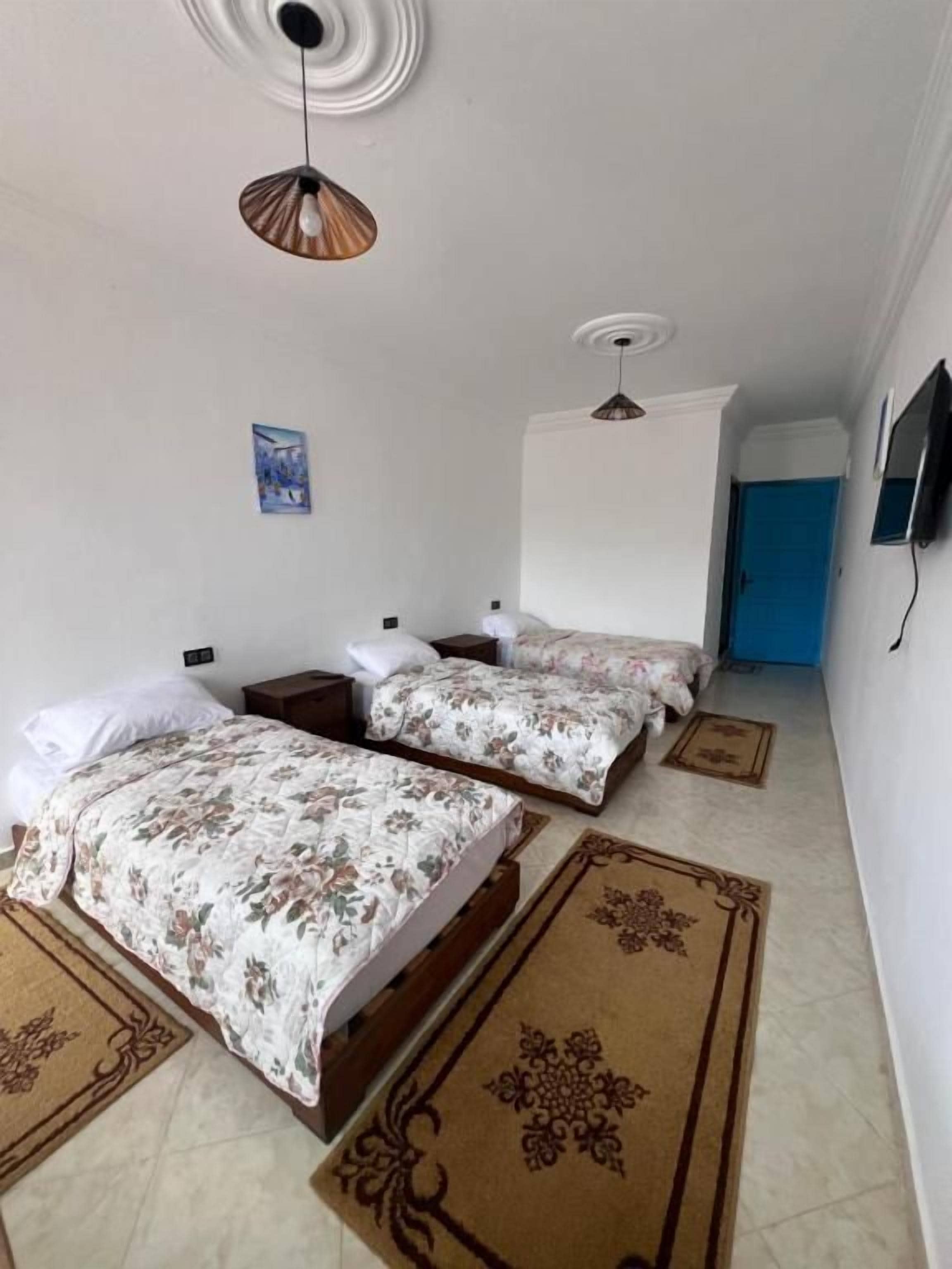 Un dormitorio con dos camas, un ventilador de techo, una puerta azul y un televisor en la pared.