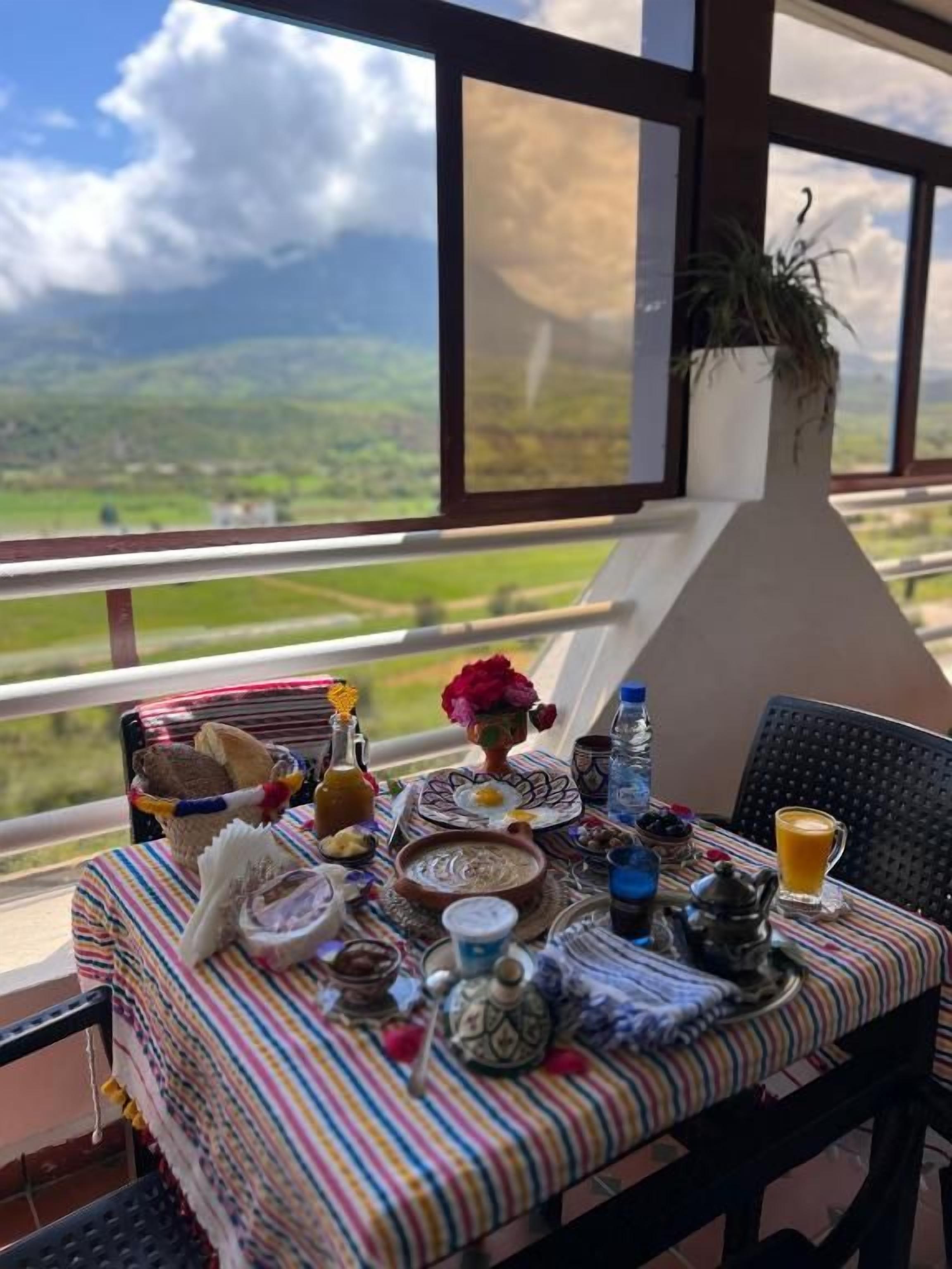 Una mesa de comedor dispuesta con diversos platos, una planta en maceta y vistas al campo a través de amplias ventanas.