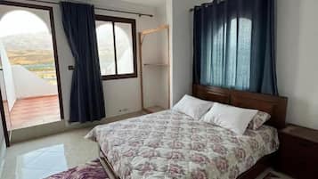 A bedroom with a bed, a nightstand, a window with curtains, and a view of the outdoors.