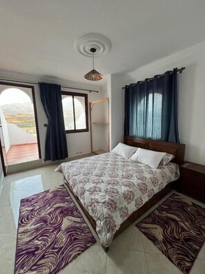 Un dormitorio con cama, mesita de noche, ventana con cortinas y vistas al exterior.
