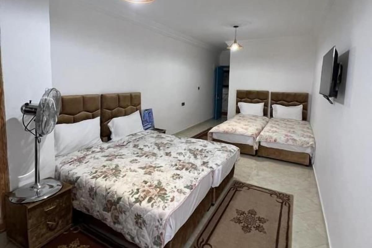 A bedroom with a large bed, two smaller beds, a ceiling fan, a TV, and a door leading to another room.