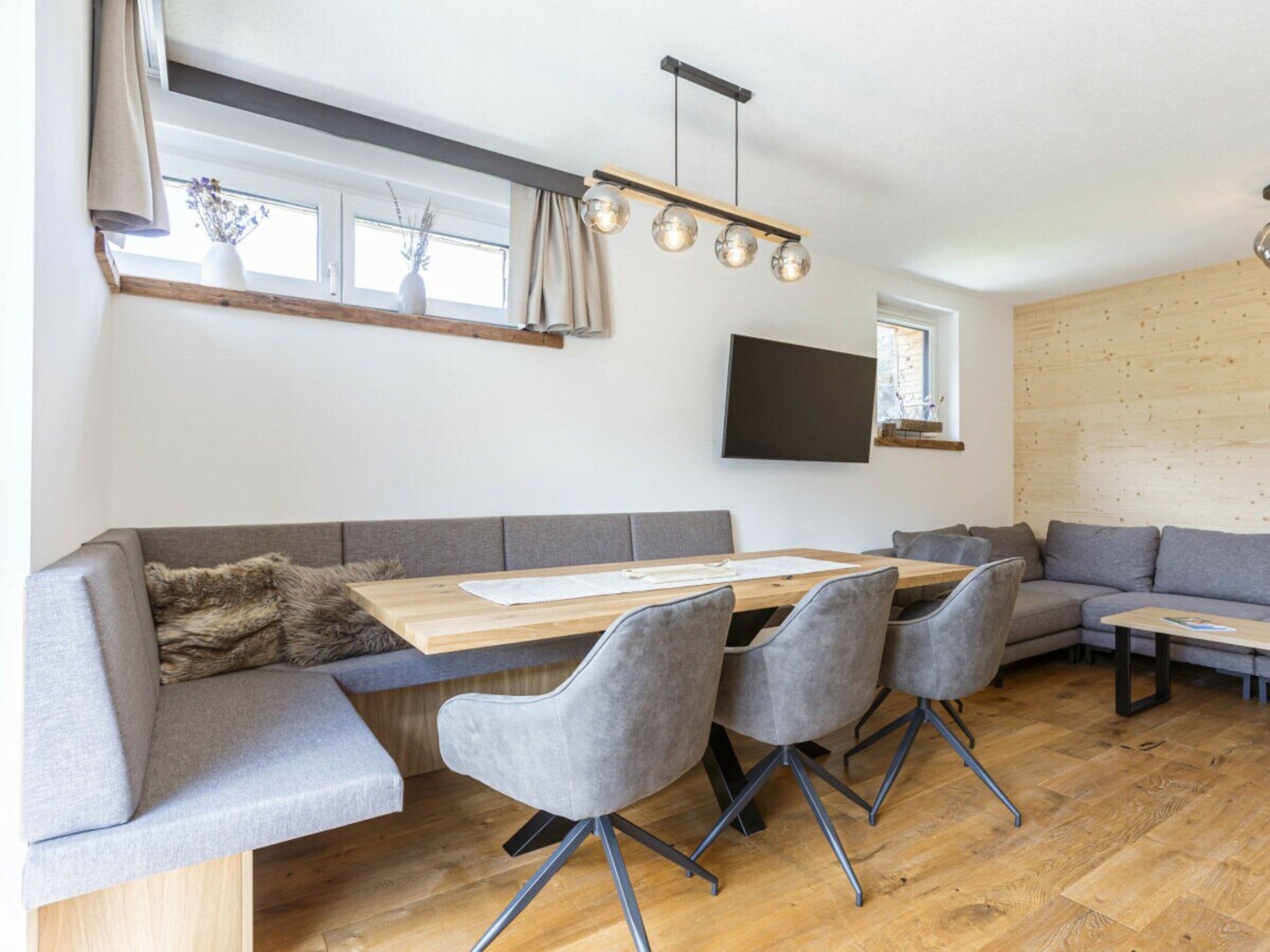 A modern living space with a wooden dining table, grey upholstered seating, and a flat-screen TV mounted on the wall.