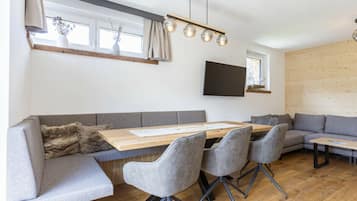 A modern living space with a wooden dining table, grey upholstered seating, and a flat-screen TV mounted on the wall.
