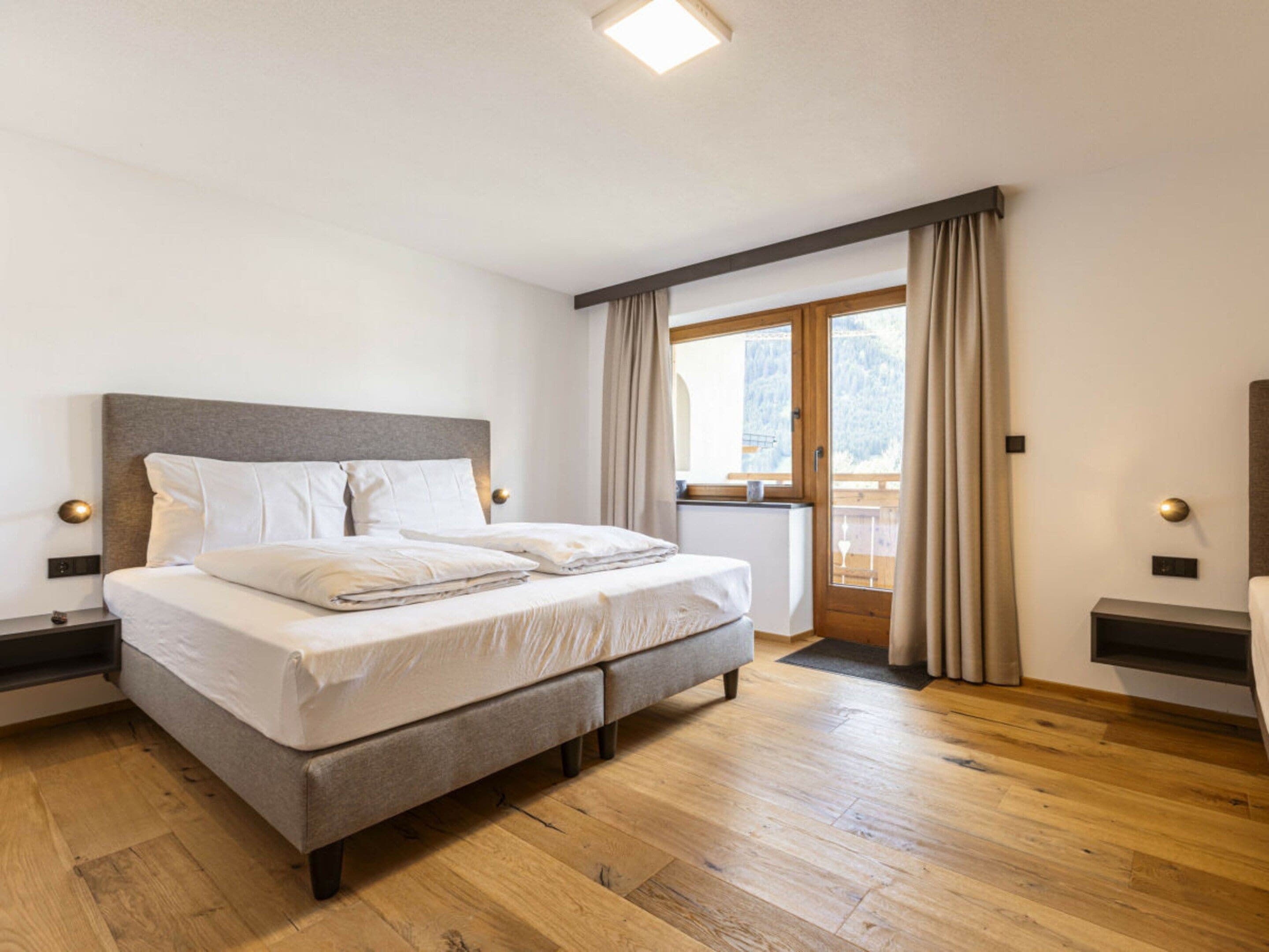 A bedroom with a bed, wooden flooring, a window with curtains, and a view of a balcony.