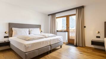 A bedroom with a bed, wooden flooring, a window with curtains, and a view of a balcony.