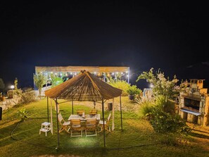 An outdoor seating area with a wooden table and chairs, surrounded by a grassy lawn and a stone structure. - Pietra & Mare Authentic Stone Villa Sea & Nature (Milas)