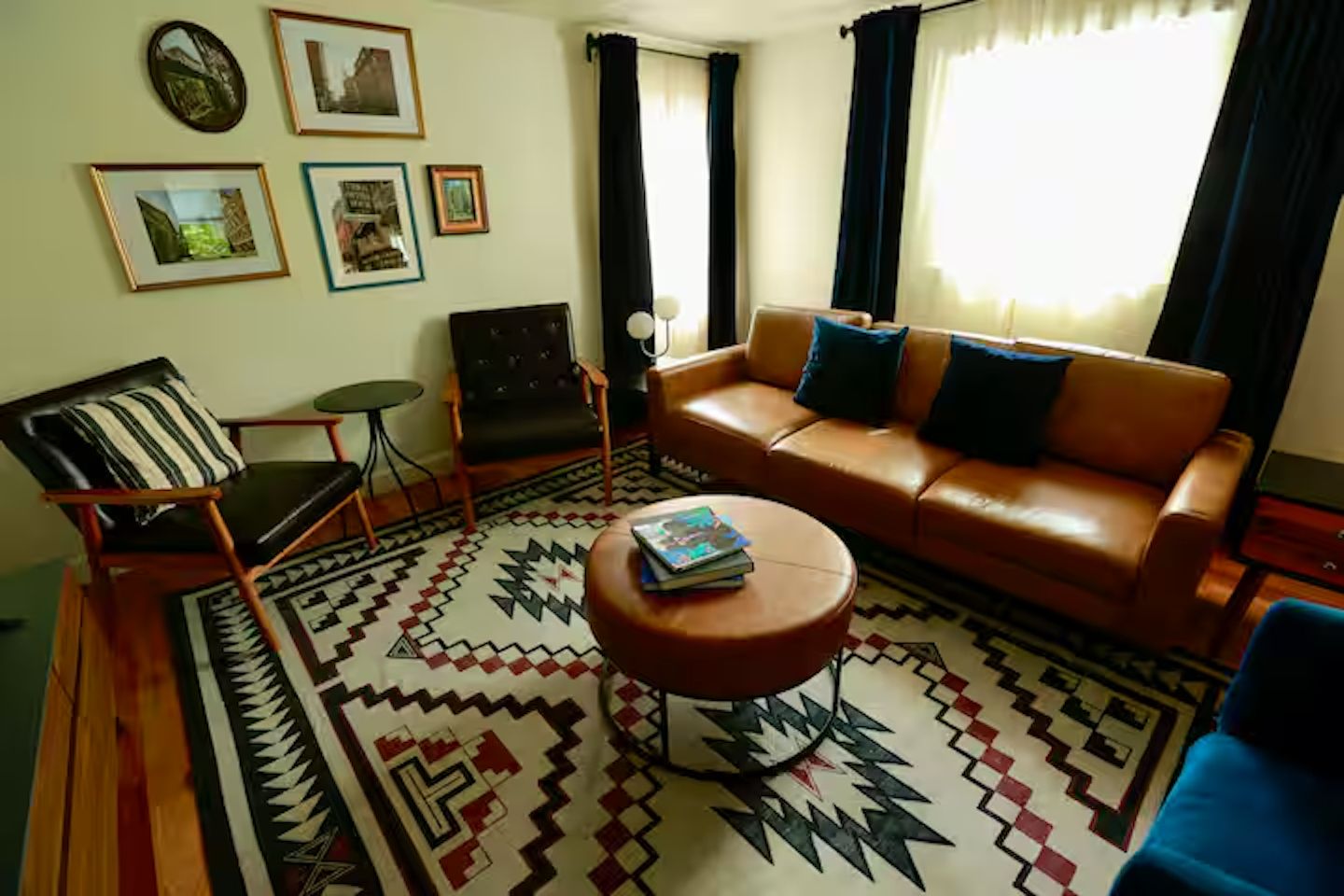 A living room with a brown leather sofa, a patterned rug, a small round table with books, a black armchair, and framed pictures on the wall.