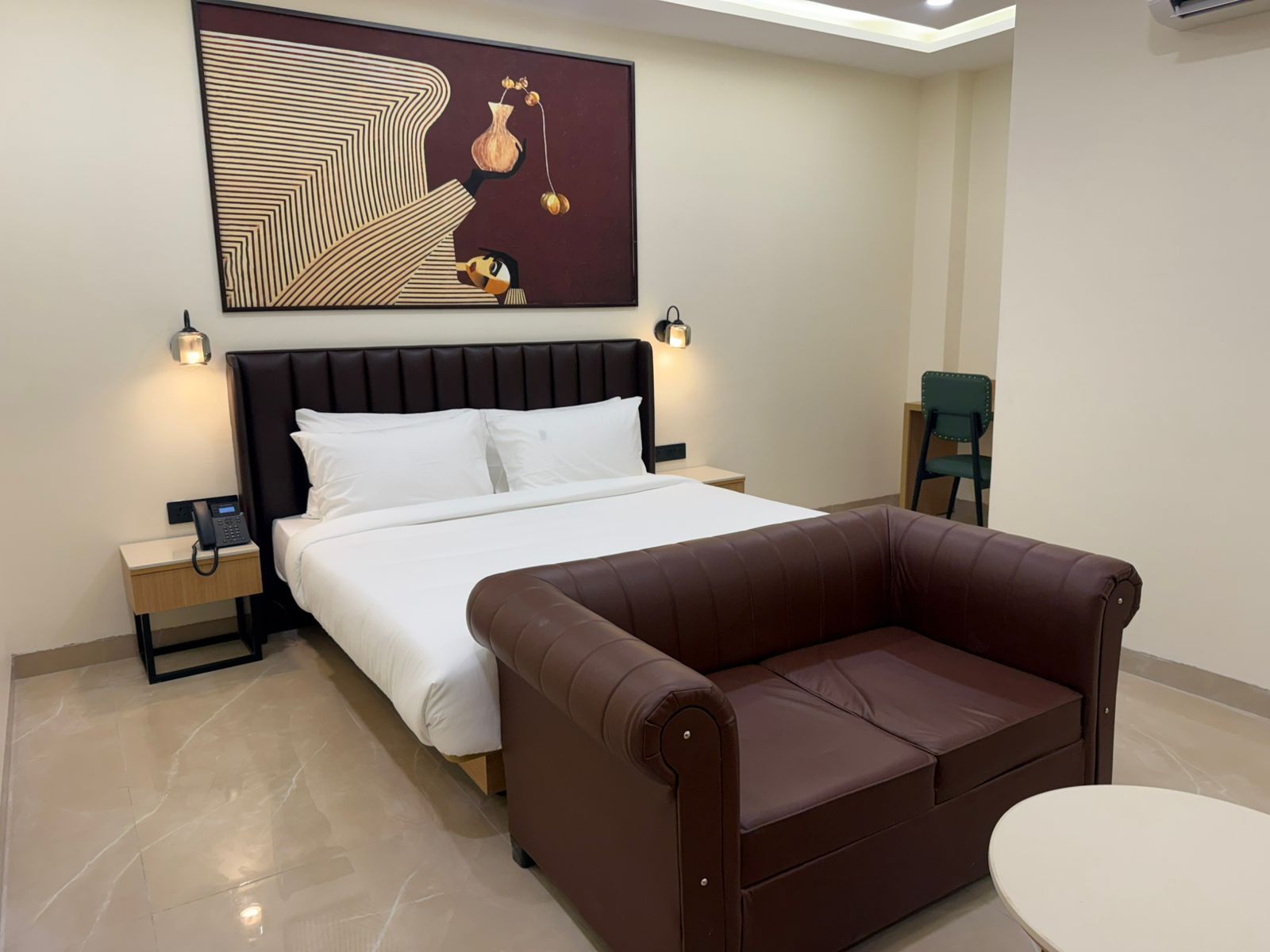 Une chambre d’hôtel comprenant un lit, un canapé en cuir marron, une petite table ronde, deux tables de chevet avec des lampes et une œuvre d’art accrochée au mur.