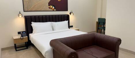 Habitación de hotel con una cama, un sofá de cuero marrón, una mesita redonda pequeña, dos mesitas de noche con lámparas y una obra de arte colgada en la pared.