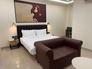 A hotel room with a bed, a brown leather sofa, a small round table, two bedside tables with lamps, and a wall-mounted artwork. - Saltstayz Premier Noida Sector 135 (Noida)